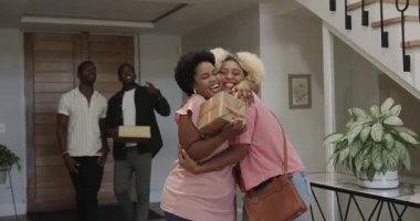 Çeşitli arkadaşlar evde kadınları selamlıyor, hediyelerini tutuyor ve içten bir şekilde gülümsüyor. Arkadaşlık, kutlama, eve dönüş, toplanma, sıcaklık, karşılama.