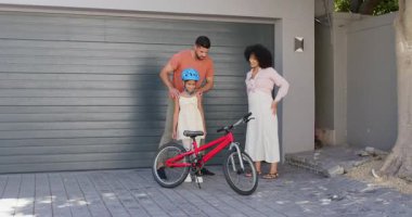 Çeşitli babalar çocukların bisiklete binmesine yardım ederken, anneleri de onları seyrediyor ve gülümsüyor. Aile, ebeveynlik, çocukluk, öğrenme, bağlanma, destek