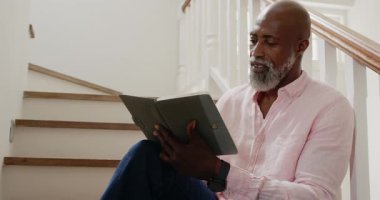 Afrikalı Amerikalı bir adam merdivenlerde kitap okuyor, evinde huzurlu bir anın tadını çıkarıyor, fotokopi çekiyor. Edebiyat, yaşam tarzı, rahatlama, sükunet, rahatlık, farkındalık.