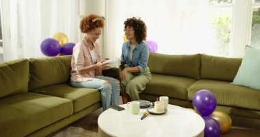 Evde kutlama yapıyorlar, neşeli lezbiyen çift koltukta hediyelerini değiştiriyorlar etrafta balonlar var. Kutlama, mutluluk, romantizm, birliktelik, sürpriz, samimi