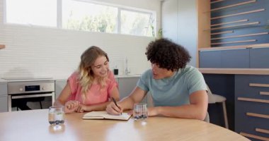 Genç çift mutfak masasında planlarını tartışıyor, birlikte defter okuyorlar, evde. Planlama, ilişki, birliktelik, sohbet, işbirliği, ev hayatı