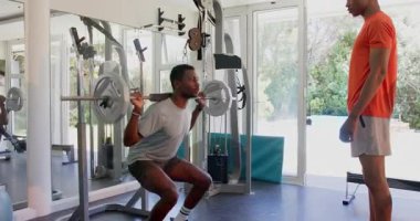 Genç adam spor salonunda ağırlık kaldırıyor. Arkadaşlarıyla antrenörlük yapıyor ve destek oluyor. Fitness, egzersiz, antrenman, egzersiz, güç, vücut geliştirme