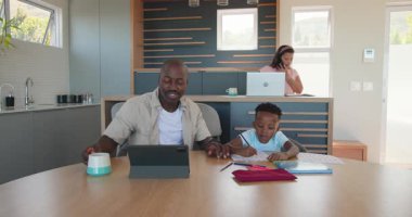 Afrika kökenli Amerikalı baba ve oğul evde tablet kullanırken çeşitli anneler yakınlarda dizüstü bilgisayarla çalışıyor. Aile, teknoloji, ebeveynlik, bağlantı, çoklu görev, aygıtlar