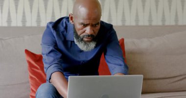 Kanepede dizüstü bilgisayara yaslanan Afro-Amerikalı adam analiz için basılı çizelgeleri daktilo ediyor, inceliyor. Profesyonel, çalışma istasyonu, modern, danışmanlık, çalışma alanı, analitik, odaklanmış