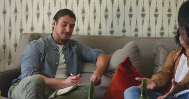 Farklı erkek arkadaşlar kanepede oturup sohbet ederken not defterine yazıp bira kızartıyorlar. İşbirliği, yoldaşlık, rahatlık, rahatlama, sohbet, kentsel, çağdaş