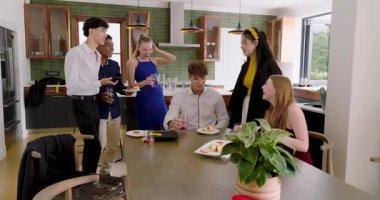 Çeşitli arkadaşlar mutfak masasına aperatif getiren denizci gömlekli adamdan sonra gülerek kadeh tokuşturuyor. Kutlama, sosyalleşme, dostluk, etkinlik, ziyafet, kahkaha, tazelik