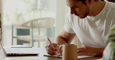 Belgeleri almak, evde çalışan bir adam, dizüstü bilgisayarın yanına yazı yazmak, kupadan yudumlamak. Konsantrasyon, üretkenlik, ev ofisi, doğal ışıklandırma, modern çalışma alanı, yansıtıcı, farkındalık