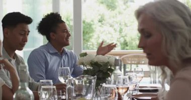 Kıvırcık saçlı bir adamın farklı arkadaşlarıyla konuşup kadeh tokuşturarak ev masasına gülmesi. Toplanma, sosyalleşme, kutlama, arkadaşlık, zarafet, boş zaman, doğal ışık.