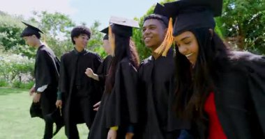 Havan topu kapaklı elbiseler giyen çeşitli mezunlar mezuniyeti kutlamak için çimlere yürüyorlar. Şenlik, başarı, yoldaşlık, açık hava, kutlama, gençlik, coşku