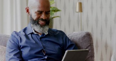 Olgun bir Afro-Amerikan adam kanepeye oturuyor, tableti tıkırdatıyor, masada çalışmak için belgeleri inceliyor. Profesyonel, çalışma alanı, modern, teknoloji, konsantrasyon, ev ofisi, gündelik
