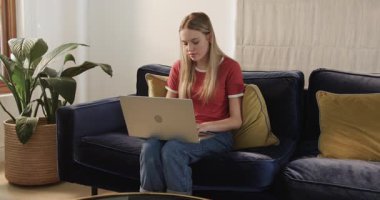 Beyaz kadın donanma kadife kanepesinde gümüş dizüstü bilgisayarda çalışıyor, akıllı telefonu seçiyor ve daktilo yazmaya devam ediyor. Rahat, çalışma alanı, modern, boş zaman, konfor, üretkenlik, ev