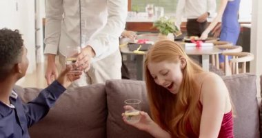 Erkek ev sahibi elinde su bardaklarıyla içeri giriyor. Oturan arkadaşlarına el çırpıyor ve gri kanepede yudumluyor. Sosyal toplanma, dostluk, modern iç mekan, gündelik kutlama, yaşam tarzı, neşeli, toplumsal