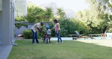 Afrikalı Amerikalı baba oğluna güneşli arka bahçede bisiklet sürmeyi öğretiyor. Çeşitli aileler, bağlılık, ebeveynlik, çocukluk, açık hava, boş zaman