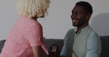 Erkek, kadının parmağına yüzük takıyor. İkisi de evde, kanepede gülümsüyor. Nişan, farklı çiftler, aşk, romantizm, mutluluk, kutlama