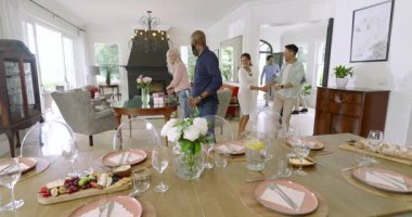 Çeşitli arkadaşlar yemek için masaya oturmadan önce yemek odasına gelip hediyeler ve şaraplar yerleştiriyorlar. Kutlama, toplanma, misafirperverlik, iç mekan, zarafet, birliktelik, sosyal