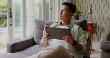 Asyalı adam kanepede cam kapılardan tablet kaydırıyor kulaklıkları çıkarıyor sallanan müzik için tıkırdatıyor. Elektronik, rahatlama, modern, yaşam tarzı, eğlence, teknoloji, ortam