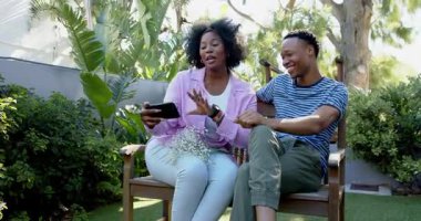 Video sohbeti, bankta oturup gülen ve bahçede selfie çeken genç bir çift. Bahçe işleri, boş zaman, açık hava, teknoloji, doğa, mutluluk.