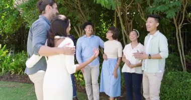 Asyalı ev sahibi kolunu uzatıyor, çeşitli aile ve arkadaşları güneşli çimlerde birlikte yürümeye davet ediyor. Açık hava toplantısı, kutlama, sosyal etkileşim, doğa, topluluk, uyum, eğlence