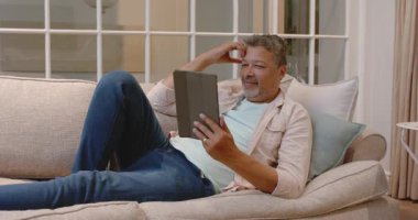 Gülümseyen kıdemli adam kanepede dinleniyor evde tablet kullanıyor, boş zamanların tadını çıkarıyor. Yaşlılar, teknoloji, rahatlama, konfor, yaşam tarzı, dijital