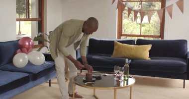 Adam evde doğum gününü kutluyor, kanepenin yanında süslerle ve pastayla dans ediyor. Kutlama, parti, eğlence, neşe, mutluluk, şenlik