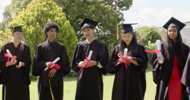 Farklı arkadaşlar çimenlikte diplomaları tutarak beşinciyi karşılıyor ve altıncı kez kollarını kavuşturup alkışlıyorlar. Kutlama, yoldaşlık, başarı, çeşitlilik, açık hava, mutluluk, birlik