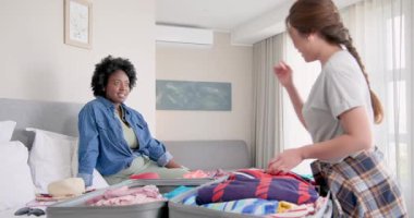Afro-Amerikalı ve Asyalı arkadaşlar, kırmızı şapkalı ve seyahat planlı modern yatak odasında bavullarını hazırlıyorlar. Tatil, arkadaşlık, seyahat, paketleme, modern, planlama, işbirliği, neşe