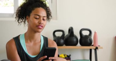 Akıllı telefonu tutan gülümseyen kadın, evde kettlebell 'lerle antrenman yaptıktan sonra dinleniyor. Fitness, teknoloji, sağlık, egzersiz, dinlenme, rahatlama