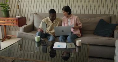 Afro-Amerikalı erkek ve Asyalı kadın kanepede çizelgeleri gözden geçiriyorlar, dizüstü bilgisayarı gösteriyorlar, kutluyorlar. İşbirliği, takım çalışması, yenilik, iletişim, üretkenlik, modern, gelişigüzel