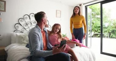 Aile yatak odasına toplanıyor, bavula havlu seriyor, baba kızına akıllı telefon gösteriyor. Aile, toplanma, ev, yaşam tarzı, rahat, rahat, samimi