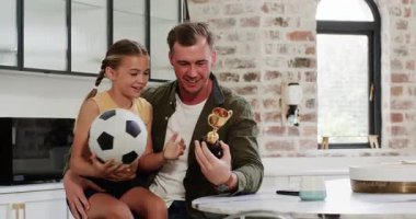 Baba ve kızı, mutfakta kupa ve top değiş tokuşu yaparak futbolu kutluyorlar. Aile, başarı, kutlama, bağlılık, gurur, başarı, gelişigüzel