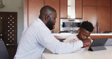 Baba oğluna mutfak masasında ödevlerinde yardım ederken anne telefonda, evde konuşuyor. Aile, ebeveynlik, çoklu görev, eğitim, yardım, ev işleri