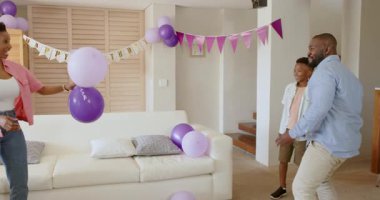 Aile evde kutlama yapıyor, mor balonlarla oynuyor ve neşeyle gülümsüyor. Kutlama, mutluluk, ev içi, eğlence, birliktelik, şakacı
