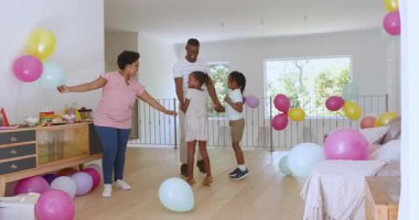 Parlak bir evde, yerde renkli balonlarla zıplayan ve dans eden Afro-Amerikan bir aile. Kutlama, neşe, balonlar, aile, coşku, dans, coşku