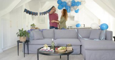 Tavan arasında doğum günü afişi keşfeden, kanepeye bakan ve kahve masasında kek düzenleyen bir çift. Kutlama, romantizm, konfor, dekor, neşe, samimiyet, ortam