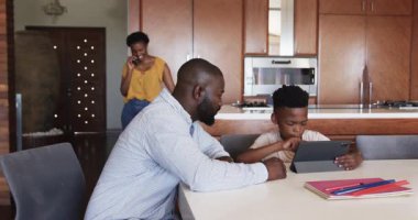 Anne evde gülümseyerek onlara katılırken baba ve oğul mutfak masasında tablet kullanıyor. Aile, bağ, teknoloji, ebeveynlik, birliktelik, iletişim