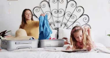 Anne bavulunu yatağın üzerine boşaltıp kıyafetlerini gösterirken kız günlük tutup kucaklıyor. Aile bağı, bakım, rahatlık, konfor, sevgi dolu, evcil, yaşam tarzı