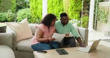 Afro-Amerikalı çift kanepeye dizüstü bilgisayar ve tabletle dokümanları tartışıyor. Toplantı, işbirliği, takım çalışması, teknoloji, evrak işleri, tartışma