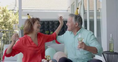 Altın şapkalı yaşlı çift terasta şarap bardaklarını tokuşturuyor, 50. pasta mumunu üflüyor, kutuyu açıyor. Kutlama, dostluk, kilometre taşı, romantizm, zarafet, boş zaman, açık hava