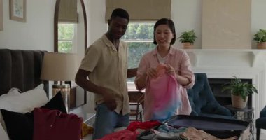 Afro-Amerikalı ve Asyalı çift yatak odasındaki açık bavulla seyahat için kıyafet ve pasaport hazırlıyorlar. Seyahat, toplanma, valiz, yerli, yaşam tarzı, günlük, örgütsel