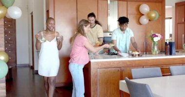 Partide, arkadaşlar mutfakta toplanıyor, yemek ve içeceklerle sofrayı kuruyor, birlikte gülümsüyorlar. Kutlama, arkadaşlık, çeşitlilik, sofra kurma, mutluluk, akşam yemeği