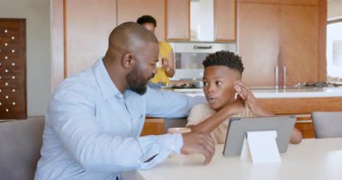 Anne mutfakta kahvaltı hazırlarken baba ve oğul tablet kullanıyor. Aile, ebeveynlik, yaşam tarzı, teknoloji, bağlanma, sabah