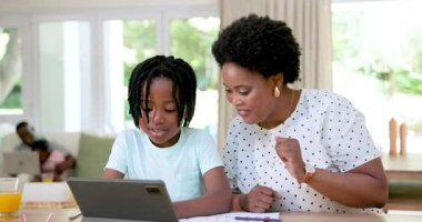 Afro-Amerikan anne ve oğlu evdeki ahşap masada oturuyor, kalemlerin yanındaki tableti işaret ediyorlar. Aile, eğitim, yaratıcılık, bağlılık, ev, etkileşim, teknoloji, doğal ışık