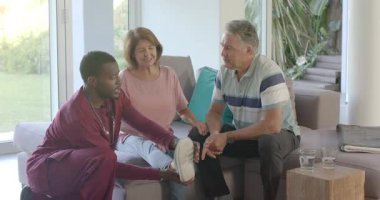 Afrikalı doktor yaşlı çifti gri koltukta selamlıyor, ayak bileğini test ediyor ve sonuçları tartışıyor. Sağlık, yaşlılık, sağlık, rehabilitasyon, ev bakımı, muayene, bakım