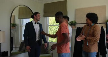 Çeşitli arkadaşlar, beyaz erkek müşterinin stüdyo aynasına geçip uygun kıyafeti giymesini izliyorlar. Moda, yoldaşlık, zanaat, stil, zarafet, takım çalışması, profesyonel