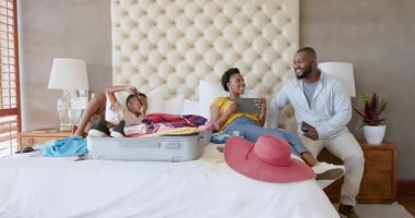 Aile bavullarını yatağa topluyor, tablet ve akıllı telefon kullanıyor, tatilin tadını çıkarıyor, evde. Seyahat, aile zamanı, teknoloji, tatil, dijital cihazlar, boş zaman