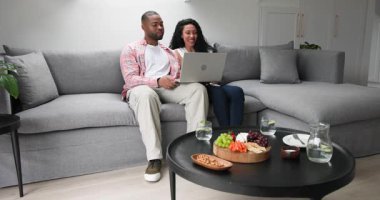 Afro-Amerikalı çift evde dizüstü bilgisayarda alarm kazanmak için tepki veriyor, aperatifleri gösterip gülüyorlar. Rahatlama, samimiyet, yerli, modern, mutluluk, rahatlık, teknoloji