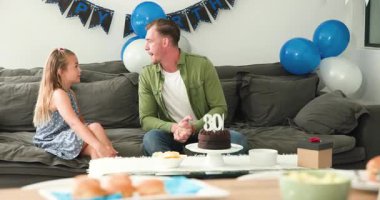 Çeşitli aileler Mutlu Yıllar afişi altında koltukta oturuyor. Anne 30. pasta için kaydırak taşıyor. Aile, kutlama, sıcaklık, birliktelik, olgunluk, bağ, neşe