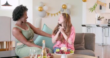 Afrikalı Amerikalı ve Asyalı arkadaşlar doğum gününü kapalı partideki kanepede pasta ve taçla kutluyorlar. Kutlama, doğum günü, arkadaşlık, konforlu, toplanma, sevinç, ikram