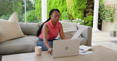 Verandada dizüstü bilgisayar kullanan Afrikalı Amerikalı bir kadın odaklanma ve kararlılıkla çalışıyor. Uzaktan çalışma, teknoloji, üretkenlik, serbest çalışma, açık hava