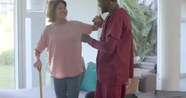 Afro-Amerikalı bakıcı yaşlı kadını baston kullanarak selamlıyor, evdeki masaya doğru adımları yönlendiriyor. Bakıcı, yaşlılar, yardım, taşınabilirlik, ev bakımı, destek, sağlık